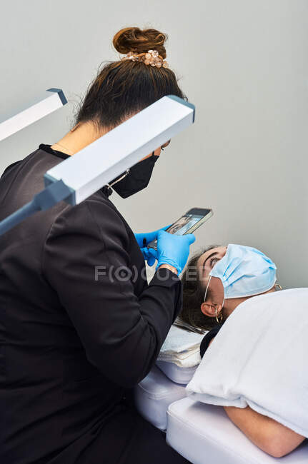 Professional cosmetologist with smartphone taking picture of face of female client getting eyelash treatment during beauty procedure in salon — Stock Photo