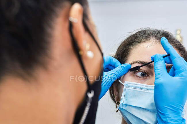 Beautician in latex gloves brushing eyebrow of young female client in protective mask during beauty appointment in modern salon — Stock Photo