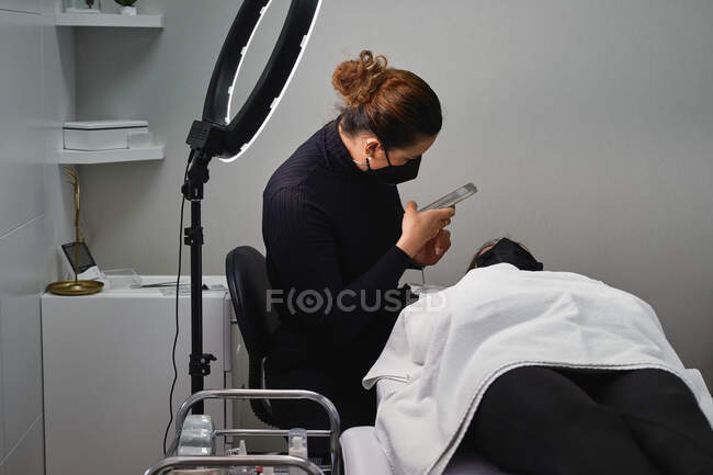 Professional cosmetologist with smartphone taking picture of face of female client getting eyelash treatment during beauty procedure in salon — Stock Photo