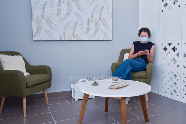 Junge Frau mit Gesichtsmaske und lässigem Outfit sitzt im Sessel und sendet Nachrichten per Mobiltelefon, während sie auf den Empfang während der Coronavirus-Pandemie wartet — Stockfoto