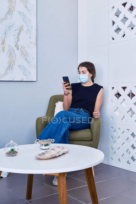Mujer joven con máscara facial y atuendo casual sentada en sillón y mensajería en el teléfono móvil mientras espera en recepción durante la pandemia de coronavirus - foto de stock