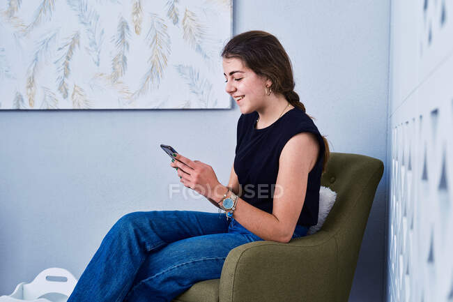 Visão lateral de positivo jovem do sexo feminino em roupa casual sentado em poltrona e mensagens no telefone móvel — Fotografia de Stock