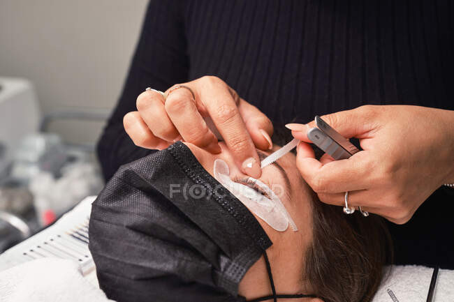 Vista laterale del cosmetico anonimo delle colture con pennello che applica vernice sulle ciglia della paziente durante la procedura di estensione delle ciglia nel salone di bellezza — Foto stock