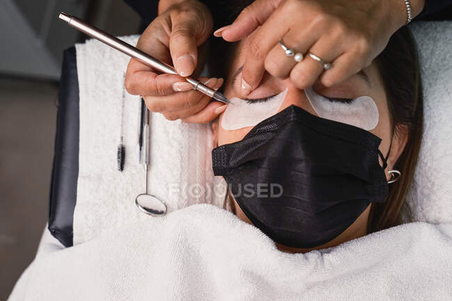 From above professional cosmetician with tweezers treating eyelashes of female customer with face mask during lash extension procedure in modern beauty salon with ring light lamp — Stock Photo