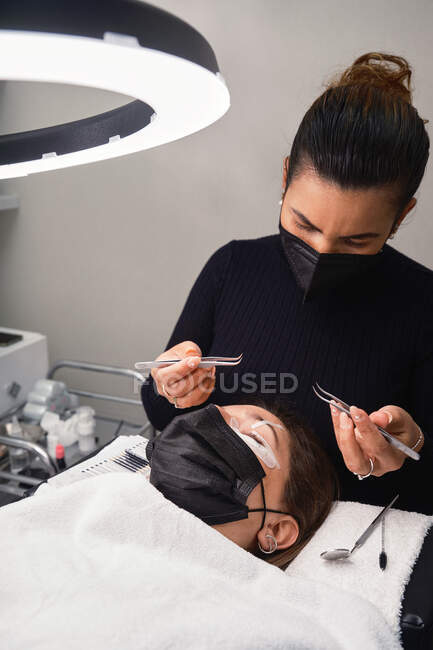 Cosmético profesional con pinzas que tratan las pestañas de cliente femenino con máscara facial durante el procedimiento de extensión de pestañas en el salón de belleza moderno con lámpara de luz de anillo - foto de stock