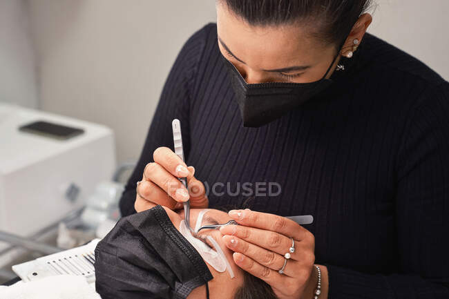 Professional cosmetician with tweezers treating eyelashes of female customer with face mask during lash extension procedure in modern beauty salon with ring light lamp — Stock Photo