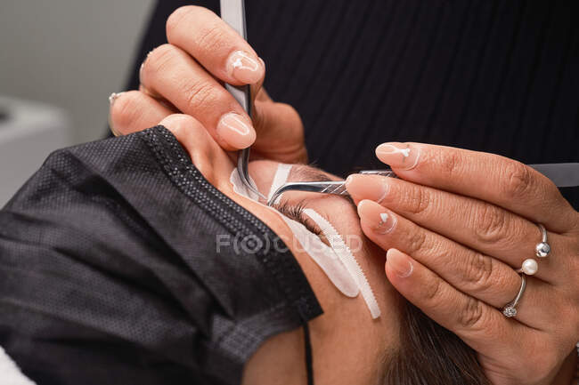 Professional cosmetician with tweezers treating eyelashes of female customer with face mask during lash extension procedure in modern beauty salon with ring light lamp — Stock Photo