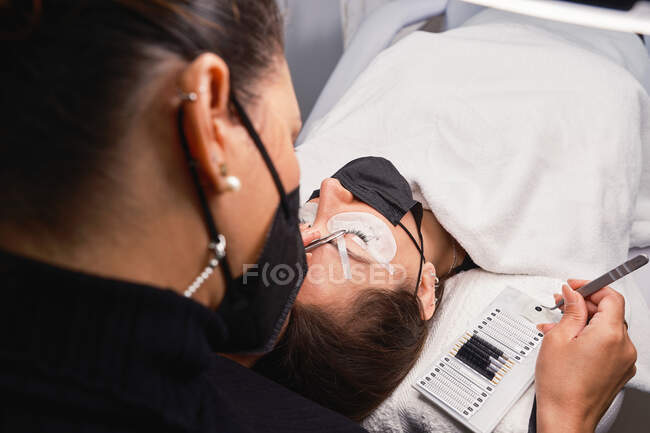 From above of professional cosmetician with tweezers treating eyelashes of female customer with face mask during lash extension procedure in modern beauty salon with ring light lamp — Stock Photo