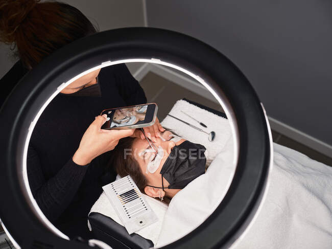 Зверху професійний косметолог з мобільним телефоном знімає процедуру розширення вій в салоні краси з кільцевим світлом — стокове фото