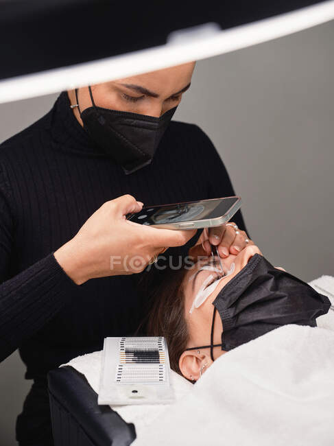 From above of professional cosmetologist with mobile phone shooting process of eyelash extension procedure in beauty salon with ring light — Stock Photo