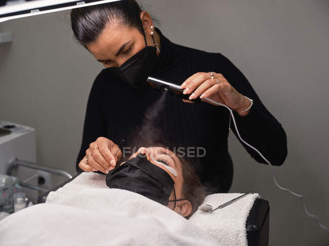 Professional beauty master using electric steamer during eyelash extension procedure to female client in salon — Stock Photo