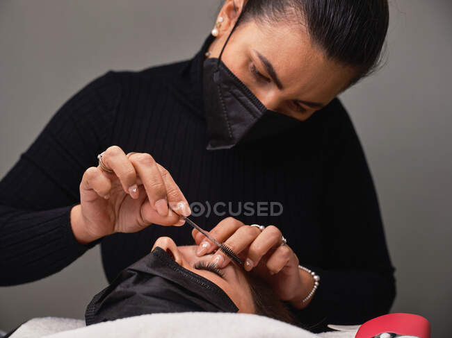 Fechar a cultura cosmetologista anônimo em luvas de látex usando pincel curler cílios durante o procedimento de beleza para o cliente feminino — Fotografia de Stock
