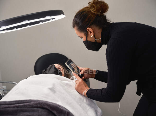 Professional cosmetologist with mobile phone shooting process of eyelash extension procedure in beauty salon with ring light — Stock Photo