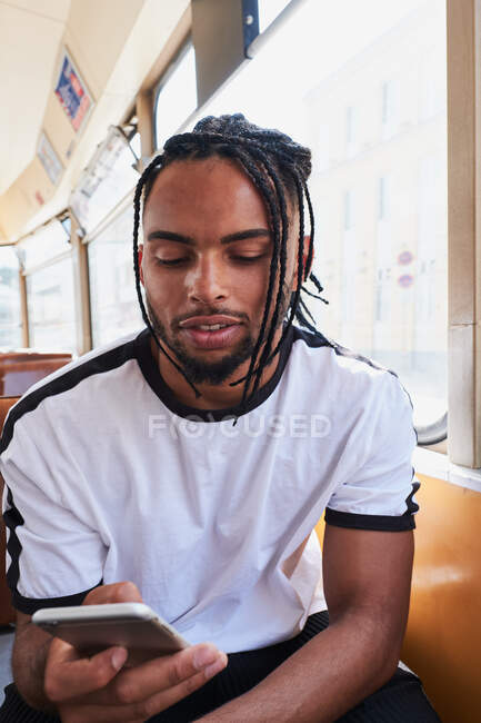 Junger positiver Afroamerikaner in lässiger Kleidung surft auf dem Handy, während er tagsüber in der Stadt im Zug auf der Viena Railway sitzt — Stockfoto