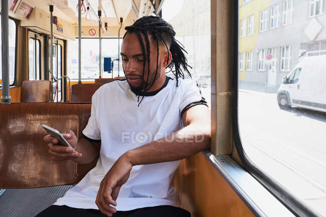 Молодий позитивний афроамериканець у повсякденному одязі, який годиться на мобільному телефоні, сидячи в поїзді на віденській залізниці вдень у місті. — стокове фото