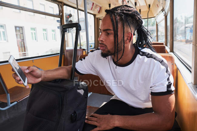 Молодий серйозний чорний чоловік у повсякденному одязі серфінг на мобільному телефоні, сидячи в трамваї з багажем під час денного світла в місті на Вієнській залізниці. — стокове фото