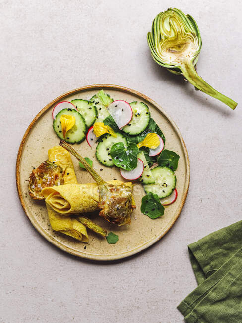 Teller von oben mit Omeletterolle und Rettich-Gurkensalat mit Kräutern in der Nähe von Artischocke und Serviette auf grauem Tisch — Stockfoto