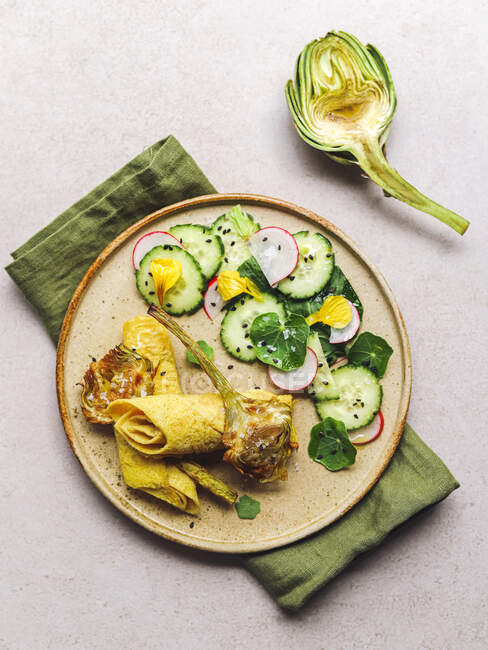 Top view of plate with omelette roll and radish and cucumber salad with herbs placed near artichoke and napkin on gray table — Stock Photo
