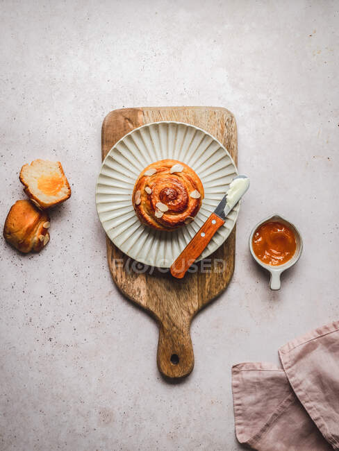 Top view of tasty cinnamon bun with almond flakes and knife with cream placed on plate on wooden cutting board near bowl with jam and napkin — Stock Photo