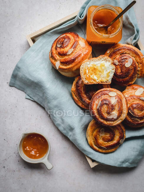 Top view of many fresh cinnamon buns with almond flakes served with jam and honey on tray covered with fabric in morning — Stock Photo