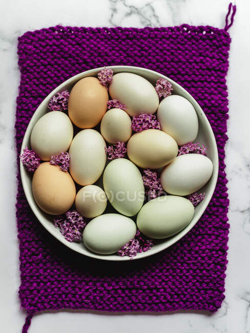 Overhead view of raw chicken eggs on round shaped plate with blossoming Lavandula flowers on purple napkin and marble surface — Stock Photo