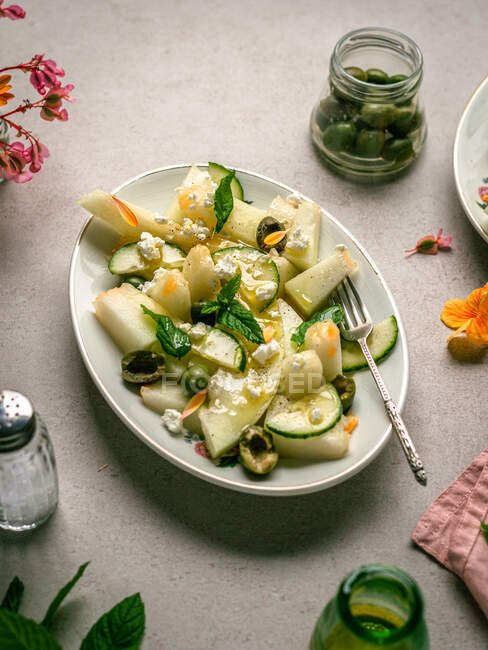 Вид на вкусный салат из дыни с огурцами и оливками подается на тарелке с травами рядом с солонкой и салфеткой — стоковое фото