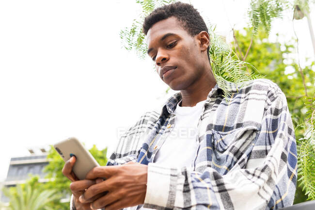 From below African American male messaging on mobile phone while standing in city street — Stock Photo