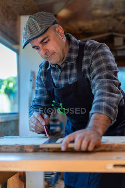 Experto adulto carpintero masculino con lápiz y regla marcando tablero de madera mientras trabaja en banco de trabajo en taller de carpintería - foto de stock