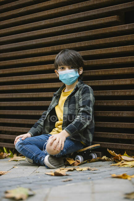 Полное тело спокойного мальчика в защитной маске сидит со скрещенными ногами у стены на скейтборде и смотрит в камеру — стоковое фото