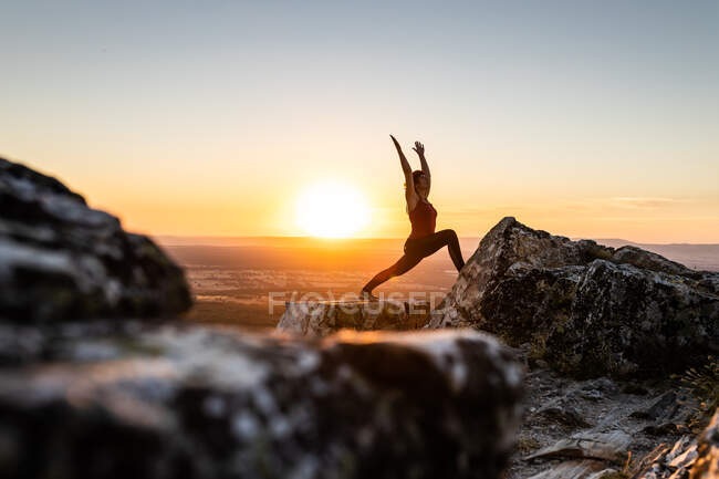 Young yogi woman practicing yoga on a rock in the mountain with the light of sunrise, side view with one leg on a rock and arms raised — Stock Photo