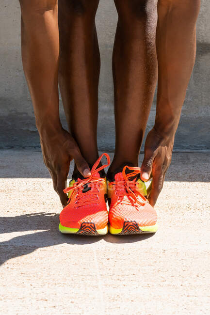 Cultivo irreconocible atleta afroamericano masculino en zapatillas de deporte vívidas haciendo doblar hacia adelante y estirar las piernas durante el entrenamiento en el día soleado en la ciudad - foto de stock
