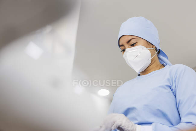 Crop giovane medico femminile in maschera sterile e cappuccio guardando giù al lavoro in ospedale — Foto stock