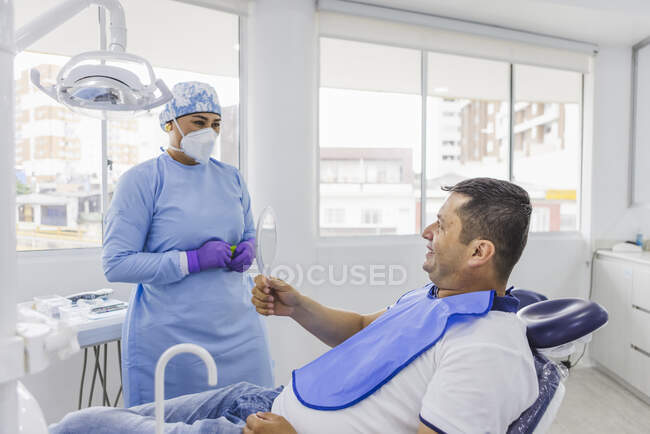 Позитивний етнічний пацієнт, який дивиться на дзеркало після процедури стоматолога в медичній формі в стоматологічному кабінеті — стокове фото