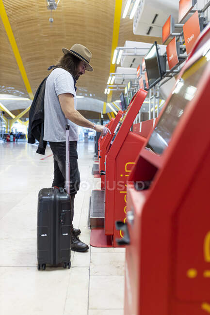 Jeune homme avec barbe et cheveux longs et chapeau achetant des billets d'avion au terminal automatique de la compagnie aérienne — Photo de stock