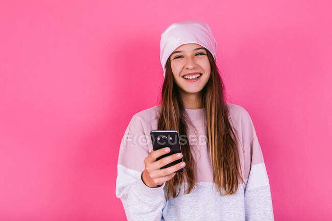 Adolescente feliz do sexo feminino com cabelo castanho no lenço de cabeça para a consciência conceito de câncer navegando no celular olhando para a câmera — Fotografia de Stock
