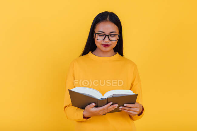 Кмітлива азійка, яка вивчає окуляри, читає підручник і готується до іспиту на жовте тло в студії — стокове фото