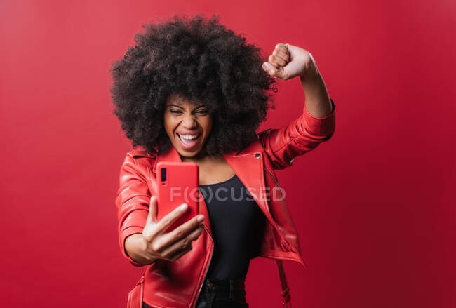 Sonriente mujer afroamericana con peinado afro tomando autorretrato en el teléfono móvil sobre fondo rojo en el estudio - foto de stock