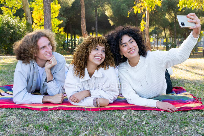 Grupo de mujeres jóvenes multirraciales y hombre con el pelo rizado acostado en la manta en el parque y tomando selfie en el teléfono móvil - foto de stock
