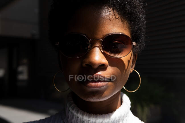 Junge Afroamerikanerin in stylischem Pullover und Sonnenbrille blickt in die Kamera, während sie im hellen Sonnenlicht vor schwarzem Hintergrund steht — Stockfoto