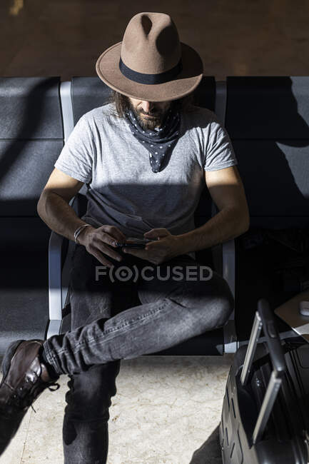Il ragazzo con il cappello in aeroporto nella sala d'attesa seduto in attesa del suo volo, con le cuffie wireless per ascoltare musica mentre chatta con il suo smartphone, vista dall'alto — Foto stock