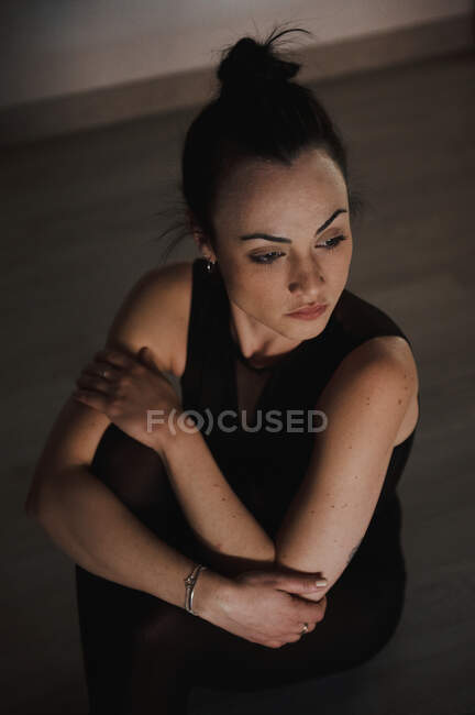 From above young tender female in sports clothes touching arms while sitting on floor and looking away — Stock Photo