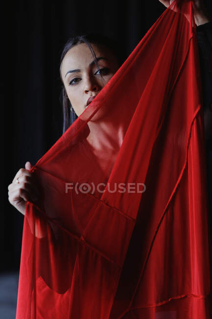 Cosecha joven hembra con labios rojos mirando a la cámara detrás de textil transparente con pliegues - foto de stock