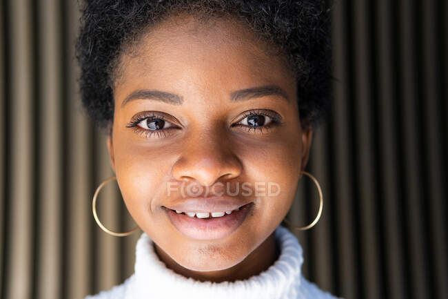 Felice elegante giovane donna nera in maglione raccolto guardando la fotocamera mentre in piedi vicino al muro edificio a strisce in città — Foto stock