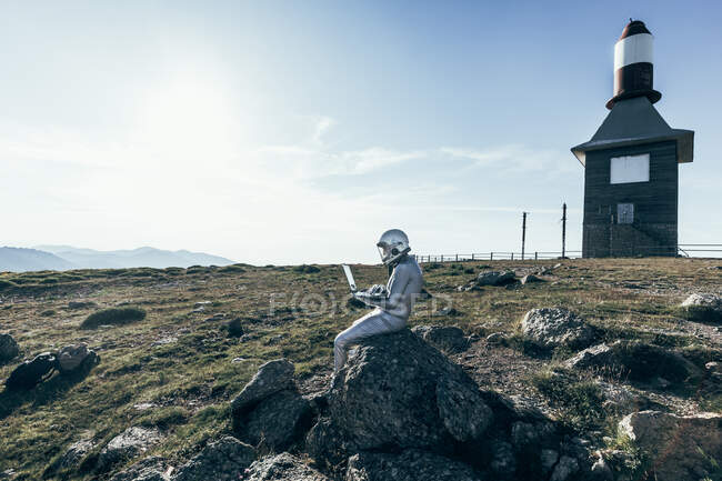 Astronauta masculino de cuerpo completo en traje espacial navegando datos en netbook mientras está sentado en la roca fuera de la estación con antenas en forma de cohete - foto de stock