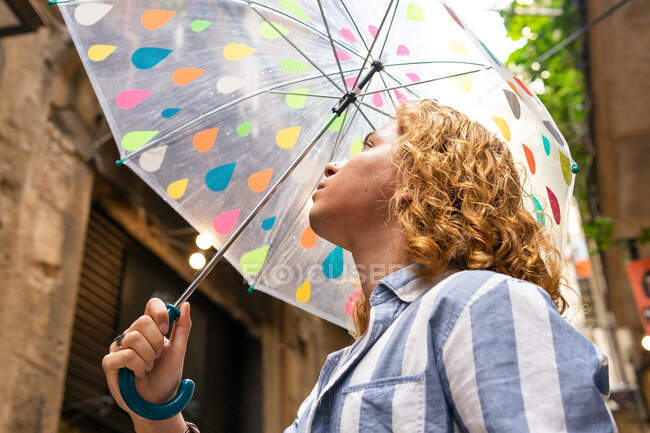 De baixo curioso macho elegante com cabelos longos em pé sob guarda-chuva transparente na rua no dia chuvoso e olhando para longe — Fotografia de Stock