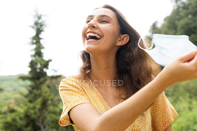 Baixo ângulo de sorrir fêmea tirando máscara médica protetora durante o coronavírus e olhando para longe na natureza no verão — Fotografia de Stock