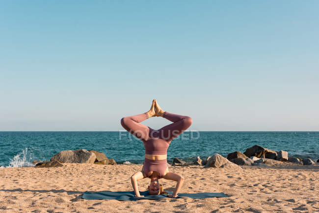 Équilibrage féminin détendu à Salamba Sirsasana sur tapis tout en faisant du yoga sur le bord de la mer — Photo de stock