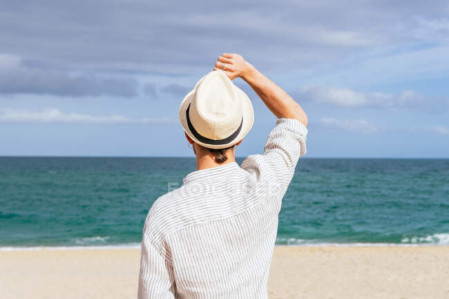 Back view of unrecognizable male in trendy casual clothes and hat walking alone on sandy beach towards waving sea while spending summer holidays on seashore — Stock Photo
