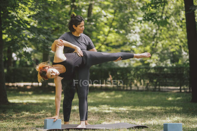Profesora de yoga apoyando a la mujer en ropa deportiva realizando la pose de Ardha Chandrasana en la esterilla durante la clase en el soleado parque - foto de stock