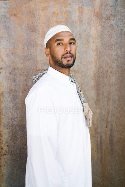 Hombre islámico en ropa blanca auténtica mirando hacia otro lado mientras está de pie contra la pared grumosa - foto de stock